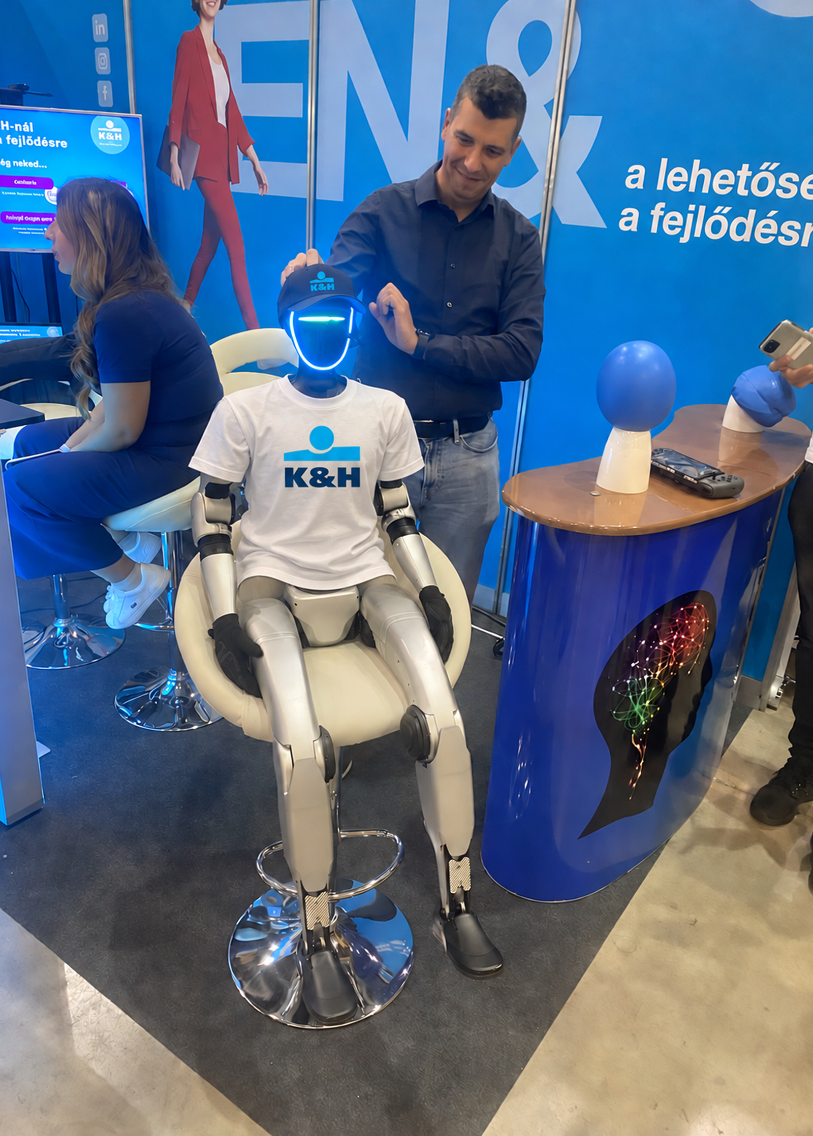 Az eventron humanoid robotja egy K&H márkajelzésű pólóban és sapkában ül a K&H rendezvénystandnál.